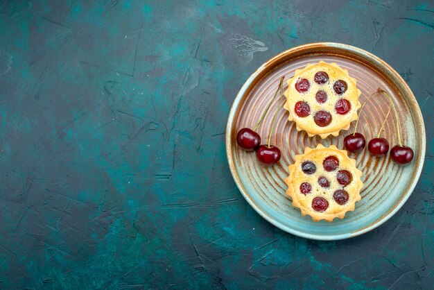 Top view of cupcakes with tasty-looking cherries on dark blue,