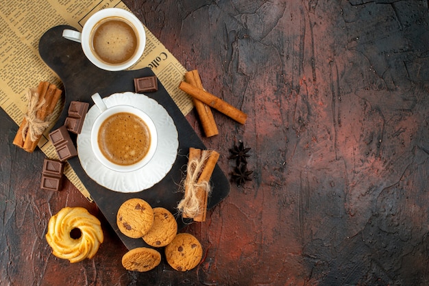 木製のまな板と古い新聞クッキーシナモンライムチョコレートバーの暗い背景の右側にコーヒーのカップの上面図
