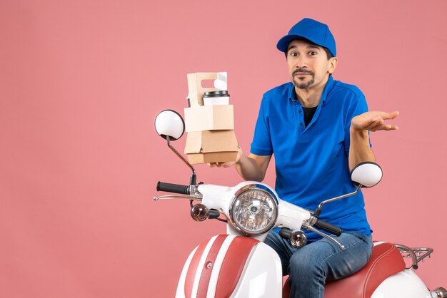 Top view of curious courier man wearing hat sitting on scooter showing orders on pastel peach background