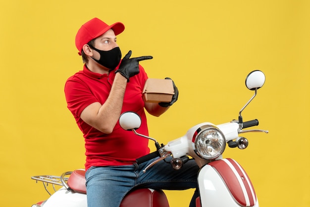 Top view of curious delivery guy wearing red blouse and hat gloves in medical mask sitting on scooter showing order