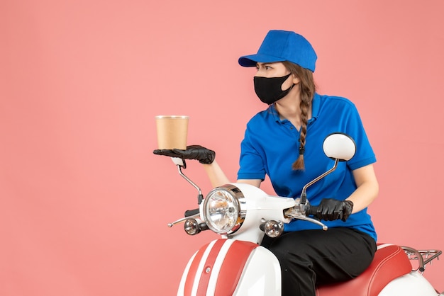 Top view of curious female courier wearing medical mask and gloves sitting on scooter delivering orders on pastel peach background