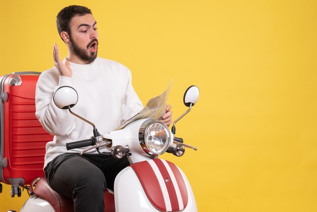 Top view of curious guy sitting on motorcycle with suitcase on it looking at map feeling confused on isolated yellow background