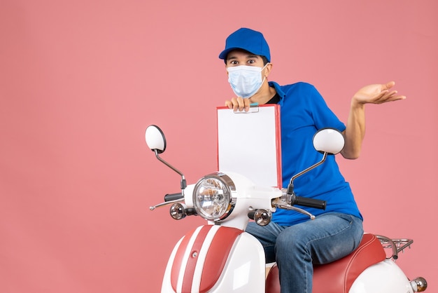 Top view of curious male delivery person in mask wearing hat sitting on scooter showing document on pastel peach background