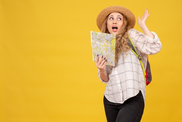 Top view of curious travelling girl wearing her hat and backpack holding map on yellow 