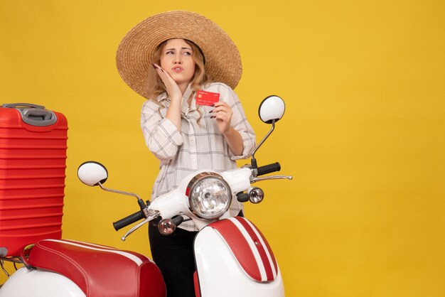 Top view of curious young woman wearing hat collecting her luggage sitting on motorcycle and holding bank card