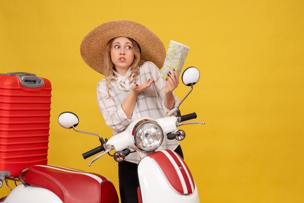 Top view of curious young woman wearing hat collecting her luggage sitting on motorcycle and holding map