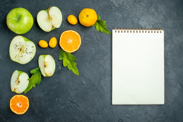 Top view cut apples and mandarines leaves cumcuat notebook on dark table