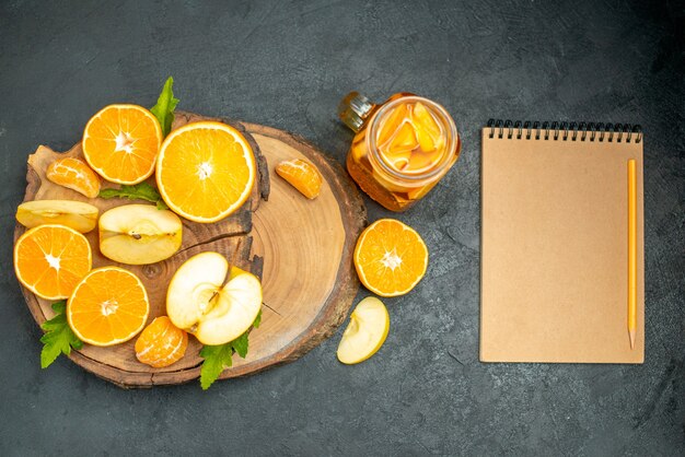 Вид сверху нарезанные яблоки и апельсины на деревянной доске, коктейль, блокнот на темном фоне