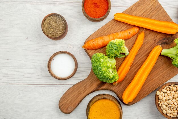 Top view cut carrots and broccoli on chopping board different spices and beans in bowls on wooden ground