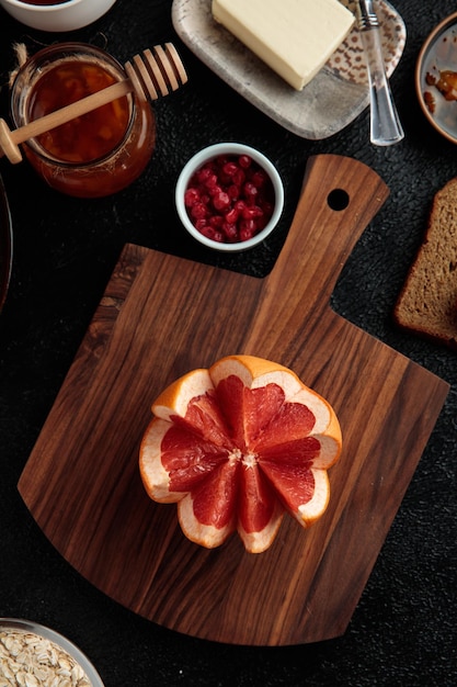 Вид сверху на нарезанный грейпфрут на разделочной доске с вареньем из красной смородины и овсяным маслом на черном фоне