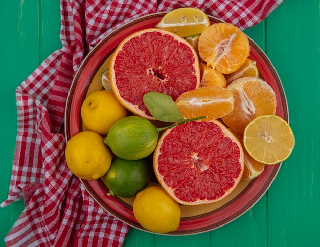 Вид сверху разрезанный пополам грейпфрут с очищенными апельсинами и лимоном с лаймом на тарелке на красном клетчатом полотенце на зеленом фоне