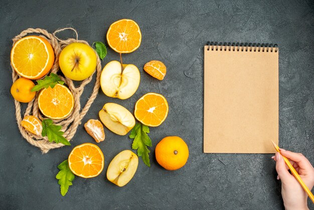 Top view cut oranges and apples a notepad pencil in female hand on dark surface