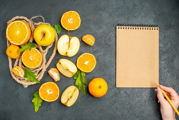 Top view cut oranges and apples a notepad pencil in female hand on dark surface