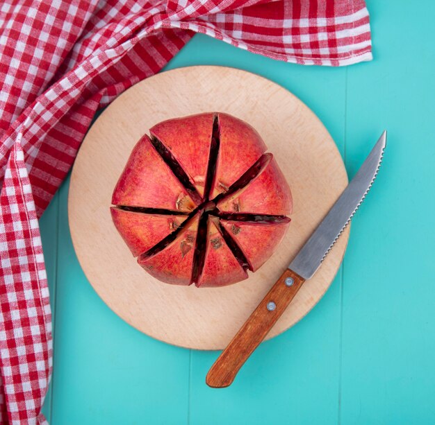 Top view of cut pomegranate on a cutting board with a knife and a red checkered towel on a blue surface