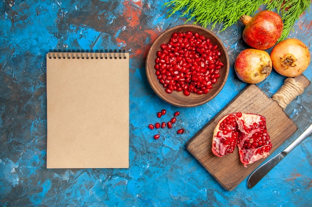 Top view a cut pomegranate and dinner knife on chopping board pomegranate seeds in bowl and pomegranates a notebook on blue background