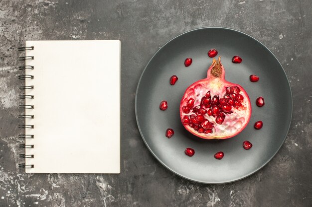 Top view cut pomegranate on oval plate a notebook on dark background