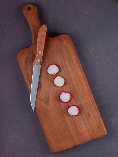 Top view of cut radishes and knife on cutting board on maroon background with copy space