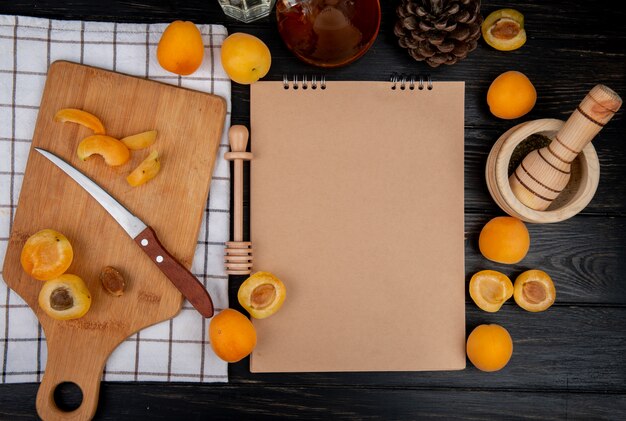 Вид сверху нарезанных и нарезанных абрикосов с ножом и блокнотом с шишкой на деревянном фоне