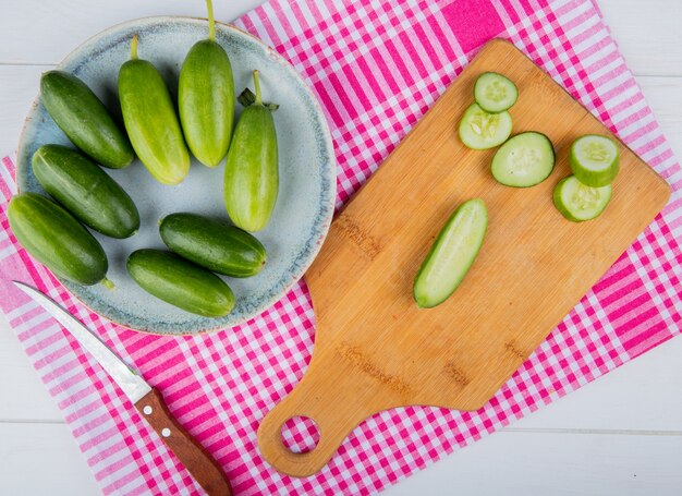 Top view of cut and sliced cucumbers on cutting board with whole ones in plate and knife on plaid cloth and wooden surface