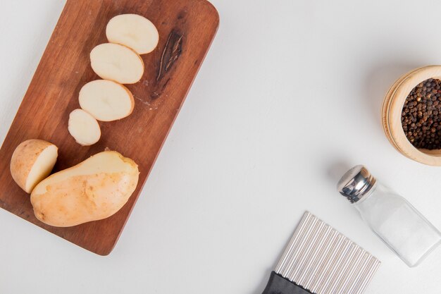 Top view of cut and sliced potato on cutting board with salt black pepper and potato chip cutter on white surface