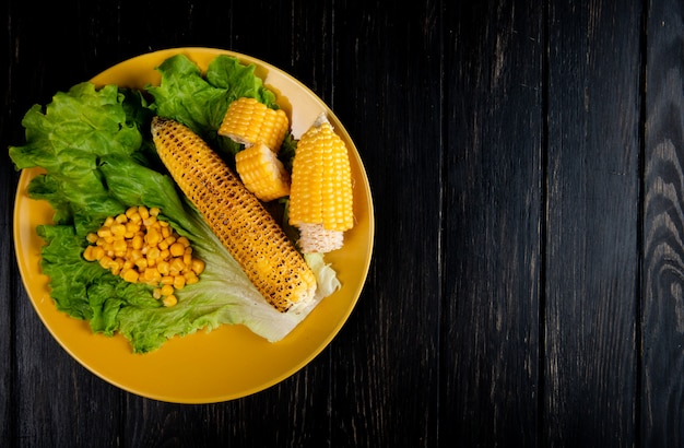 Top view of cut and whole corns and corn seeds with lettuce in plate on left side and black surface with copy space