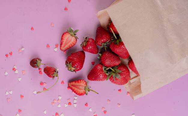 top view of cut and whole strawberries spilling out of paper bag on purple table