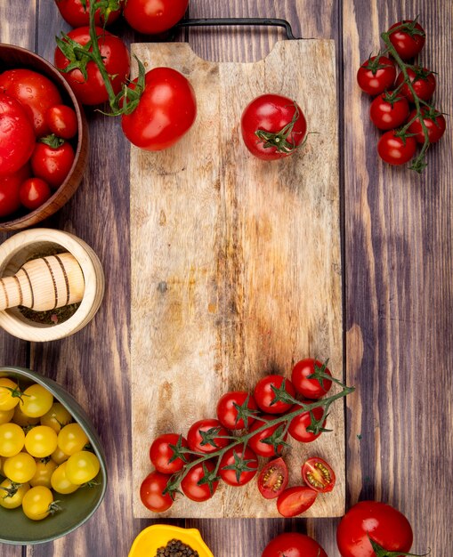 Top view of cut and whole tomatoes on cutting board with other ones black pepper garlic crusher on wooden surface