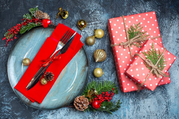 Top view of cutlery set with red ribbon on a decorative napkin on a blue plate and christmas accessories next to red gift boxes on dark background