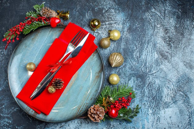 Top view of cutlery set with red ribbon on a decorative napkin on a blue plate and christmas accessories on the right sideon dark background