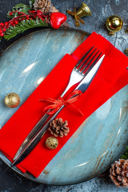 Top view of cutlery set with red ribbon and decorative napkin on blue plate decorative accessories conifer cone on dark background