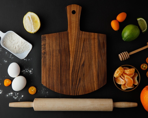 Top view of cutting board with egg lemon lime orange kumquat tangerine flour with honey dipper and rolling pin on black background