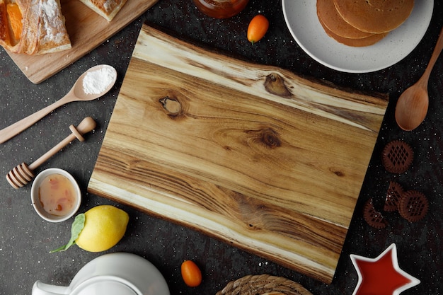 Top view of cutting board with pancakes jam lemon kumquats croissant flour cookies spoon on black background