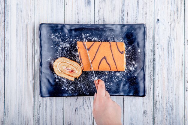 Top view of cutting a swiss roll with whipped cream and raspberry jam on black board on rustic background