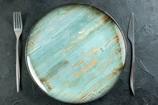 Вид сверху голубая круглая тарелка, вилка и нож на темной поверхности
