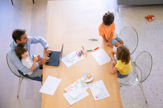 Top view of dad and kids sitting at table together. Brother and sister painting doodles with colorful pens. Middle-aged dad using laptop and holding little son. Childhood, weekend and family concept