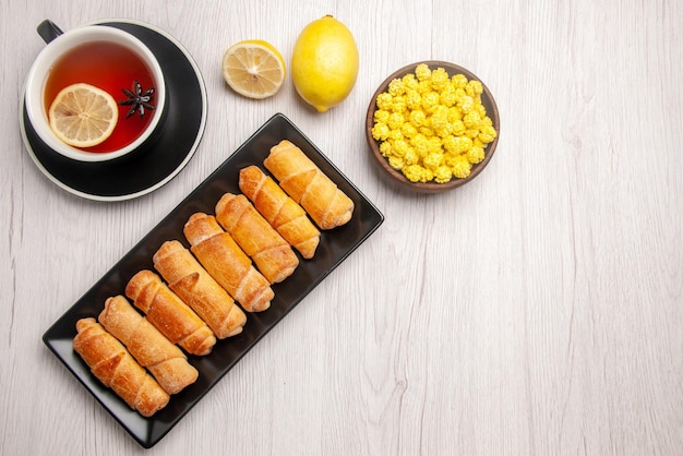 Top view dark plate of dessert lemon bowl of sweets and a cup of tea with lemon next to the dark plate of tubular pastry on the white table