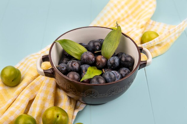 Top view of dark purple sloes on a bowl with leaves on a yellow checked tablecloth on a blue background