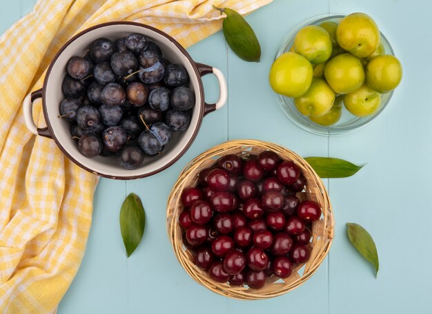 Top view of dark purple sloes on a bowl with red cherries on a bucket with green cherry plums on a blue background