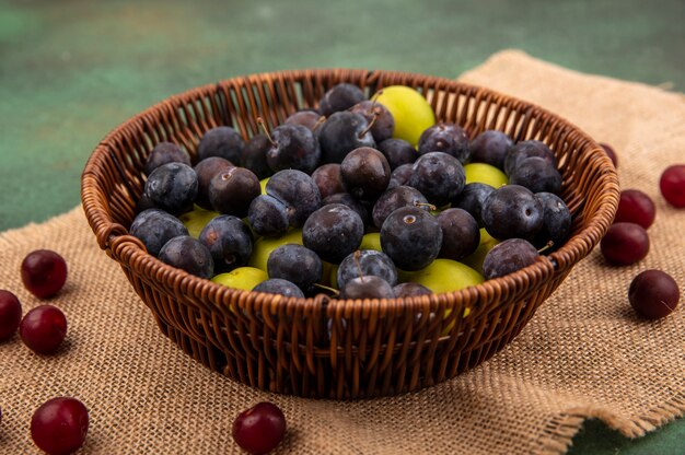 Top view of dark purple sloes on a bucket with green cherry plum on a bucket on a sack cloth on a green background