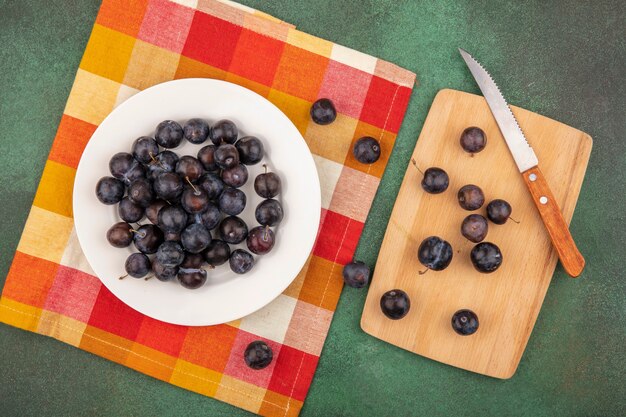 Top view of dark purple sloes on a white plate on a checked tablecloth with sloes isolated on a wooden kitchen board with knife on a green background