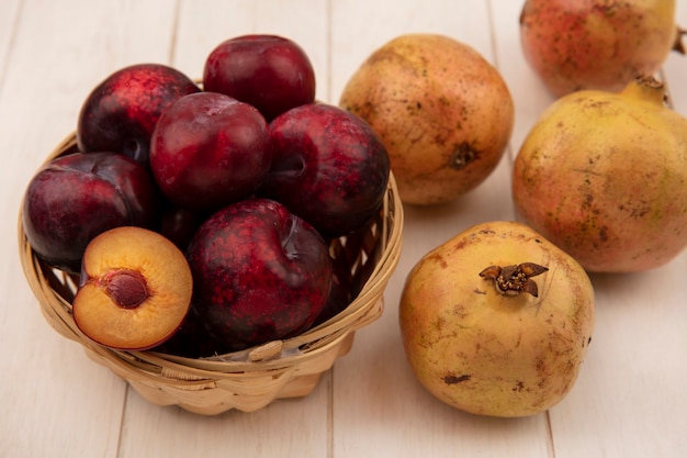 Top view of dark red skinned pluots on a bucket with pomegranates isolated on a white wooden surface