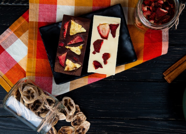 Top view of dark and white chocolate bars on a black tray with various dried sliced fruits in glass jars on black background with copy space