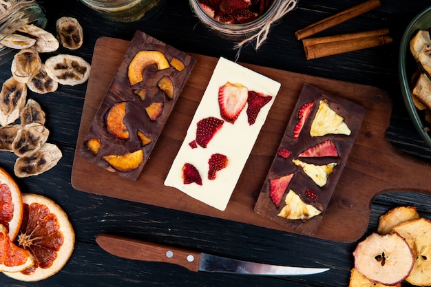 Top view of dark and white chocolate bars on a wooden cutting board with various dried sliced fruits on black background