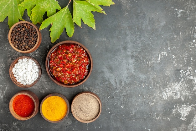 Вид сверху вкусной аджики в деревянной миске с разными специями в мисках на сером фоне