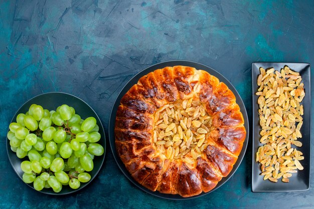 Top view delicious baked cake with raisings and fresh green grapes on dark-blue floor cake pie sugar sweet biscuit dough