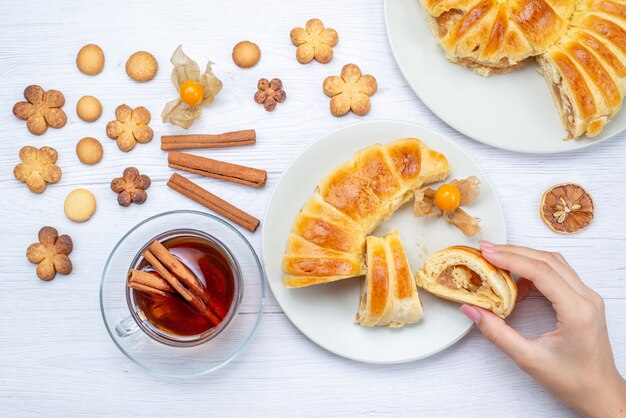 top view of delicious baked pastry with sweet filling sliced and whole along with cookies and tea on light desk, cookie biscuit pastry cake sweet tea