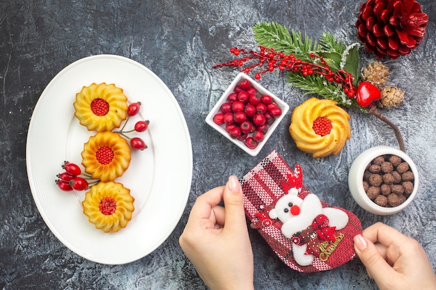 Вид сверху вкусного печенья и кизила на белой тарелке новогоднего носка с красной хвойной шишкой на темной поверхности