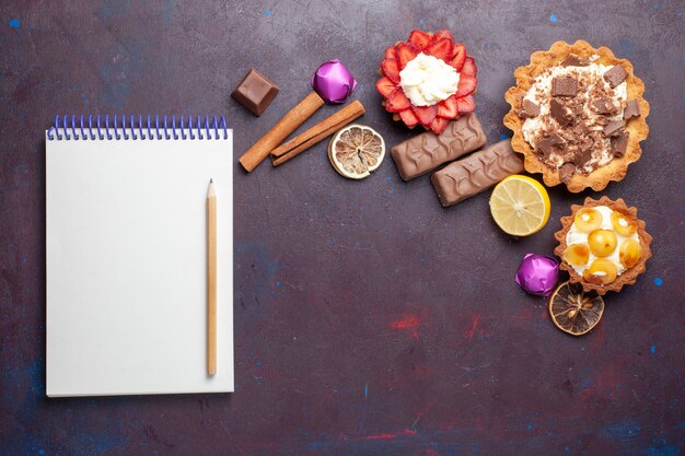 Top view of delicious cakes along with cinnamon notepad and candies on the dark surface