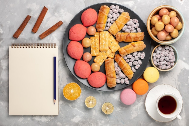 Вид сверху вкусные торты с рогаликами, крекерами, конфетами и чаем на светло-белом столе, бисквитном печенье, сладком сахарном пироге
