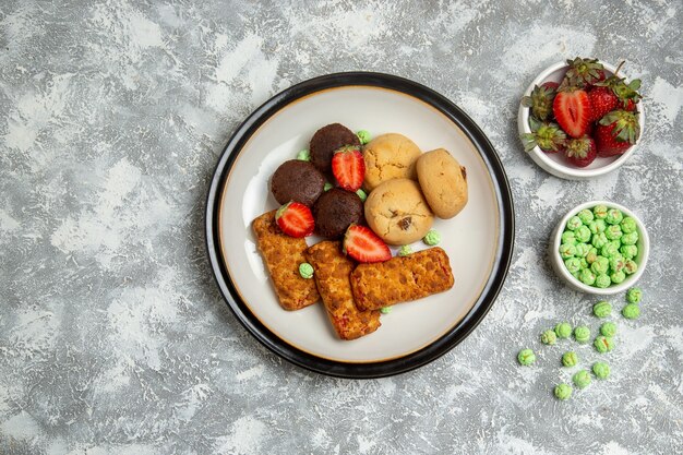 Вид сверху вкусные торты с печеньем, конфетами и клубникой на белом фоне, бисквитный сахарный торт, сладкое чайное печенье